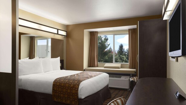 A hotel room with a bed, a desk, a chair, a TV, a window with curtains, and a view of trees. - Microtel Inn & Suites Odessa TX (Odessa)