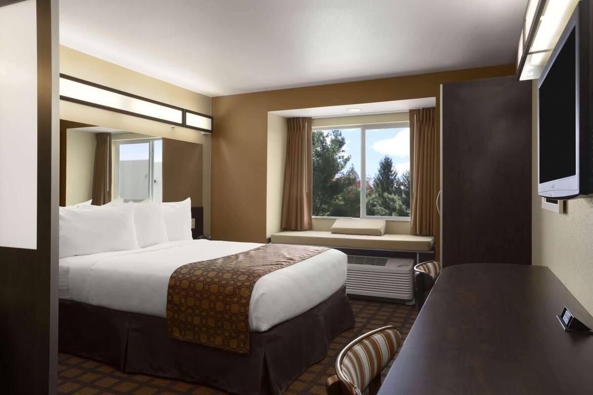 A hotel room with a bed, a desk, a chair, a TV, a window with curtains, and a view of trees.
