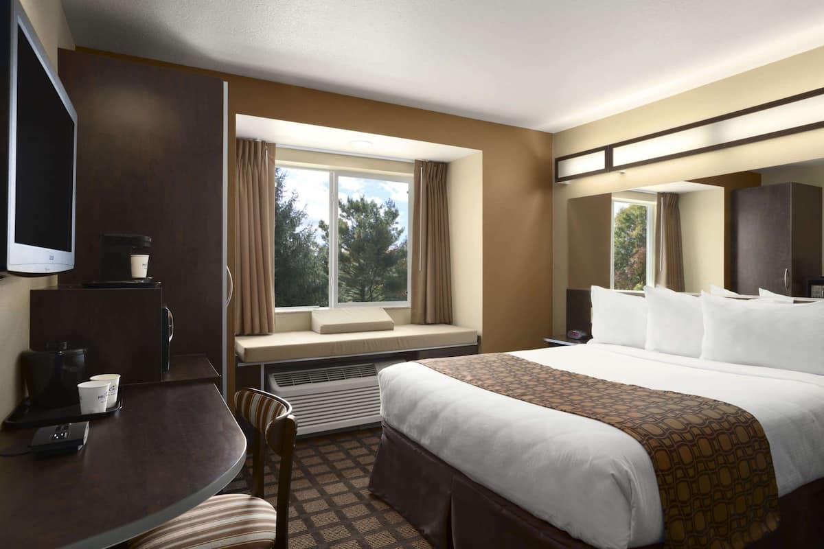 A hotel room with a bed, a desk with a coffee maker, a chair, a TV, and a window with curtains.