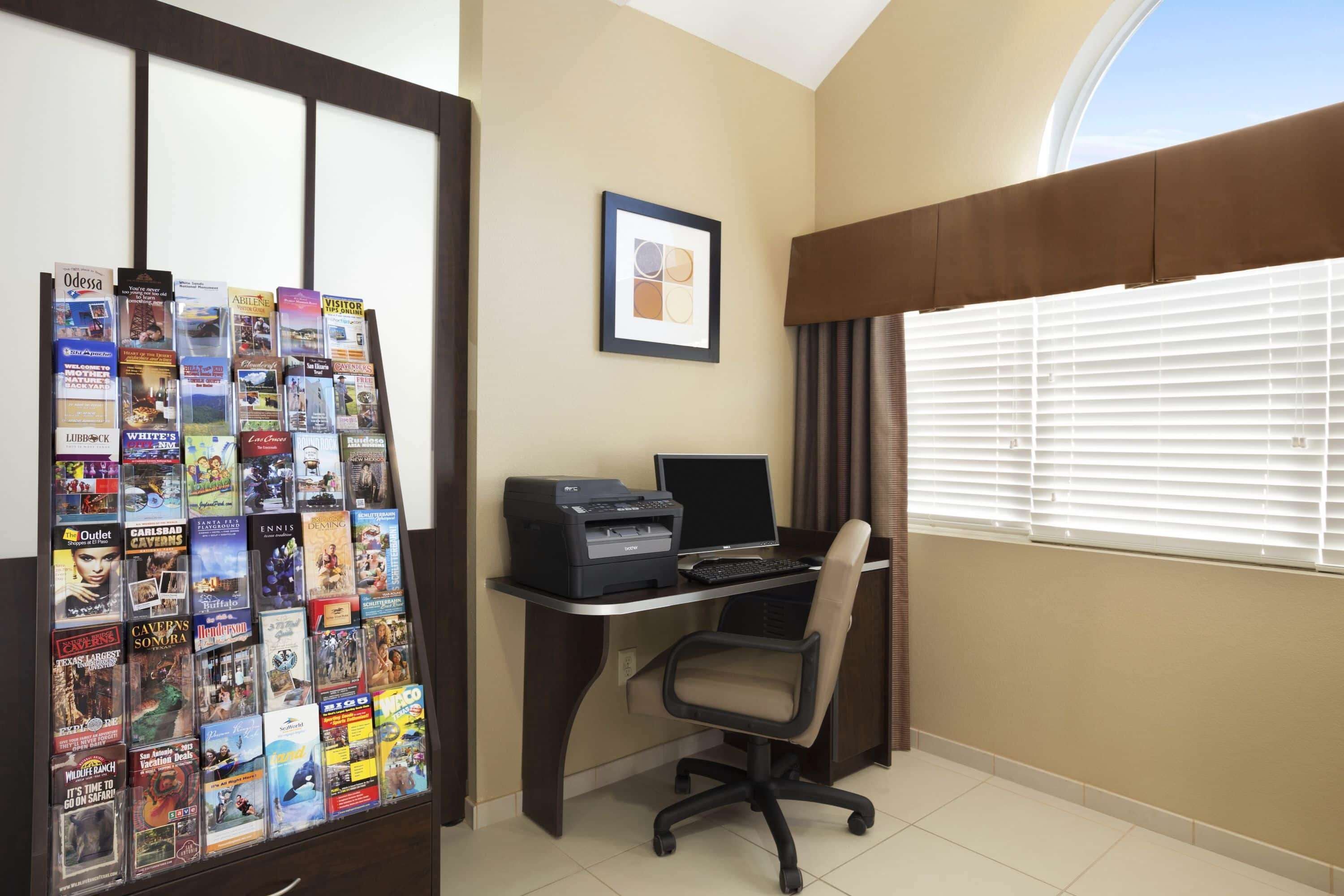 A hotel room with a desk, a printer, a chair, and a bulletin board with travel brochures.
