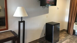 A room with a flat-screen TV mounted on the wall, a standing lamp, a small black refrigerator, and a wooden table. - Bellflower Travel Inn (Bellflower)