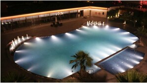 Une grande piscine extérieure dotée de plusieurs fontaines, entourée de palmiers et bordée d’un bâtiment à la façade en verre.