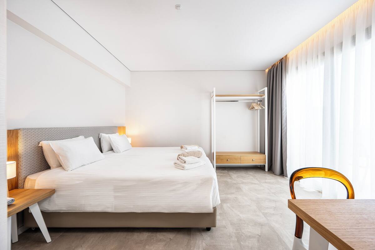 A modern hotel room with a neatly made bed, a wooden nightstand, a wooden chair, and a walk-in closet with a mirror and hanging clothes.