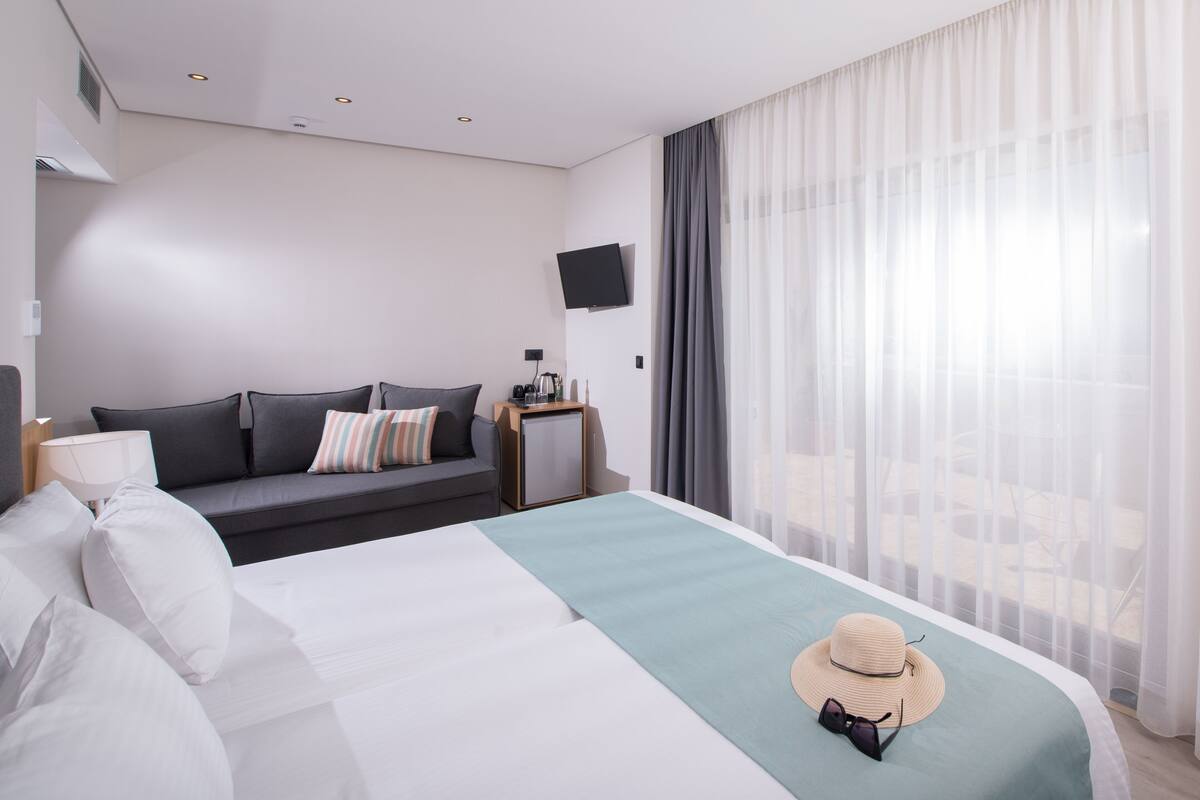 A modern hotel room with a large bed, a sofa, a TV mounted on the wall, and a view of the outdoors through sheer curtains.