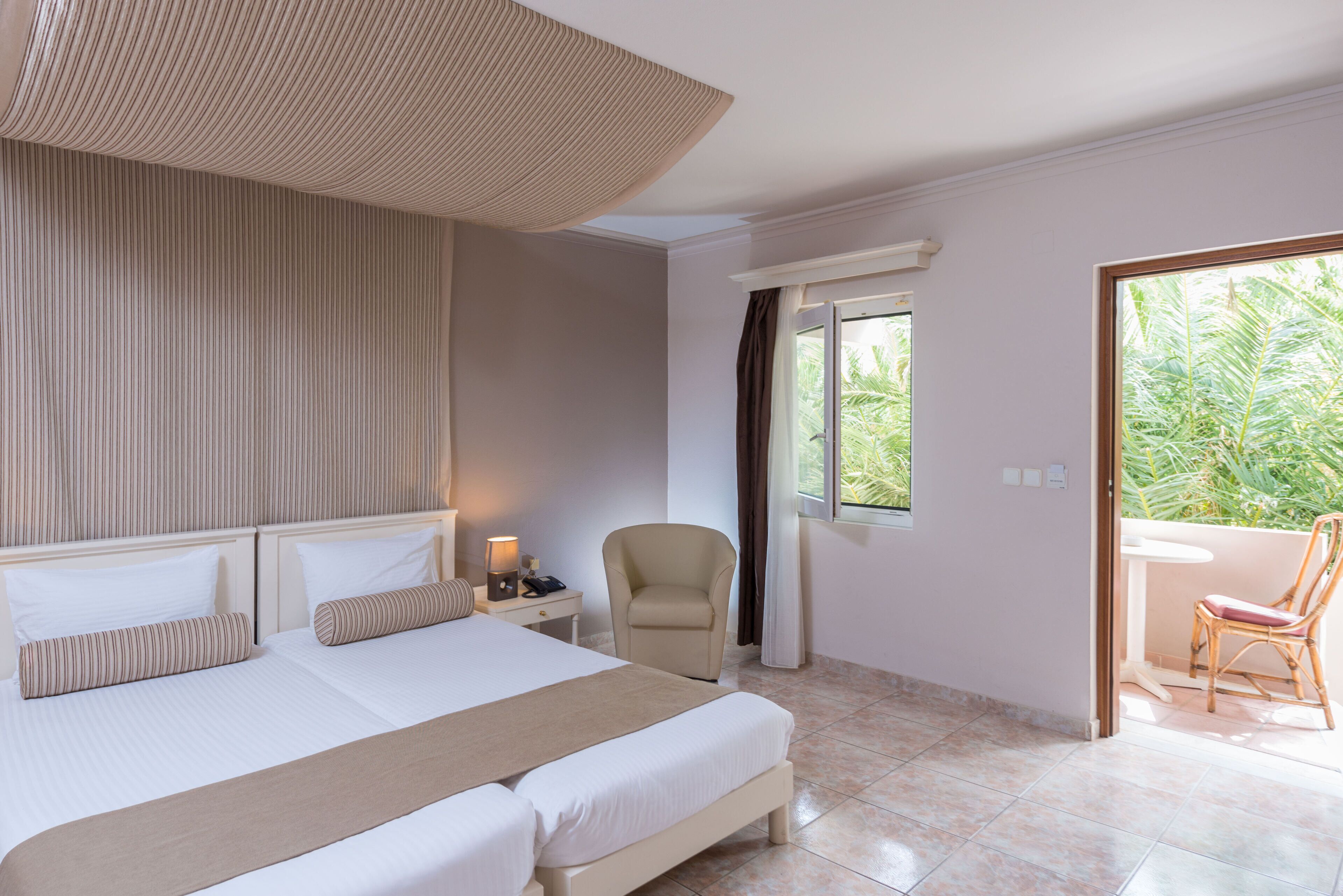 A hotel room with a large bed, a chair, a small table, and a view of greenery outside.