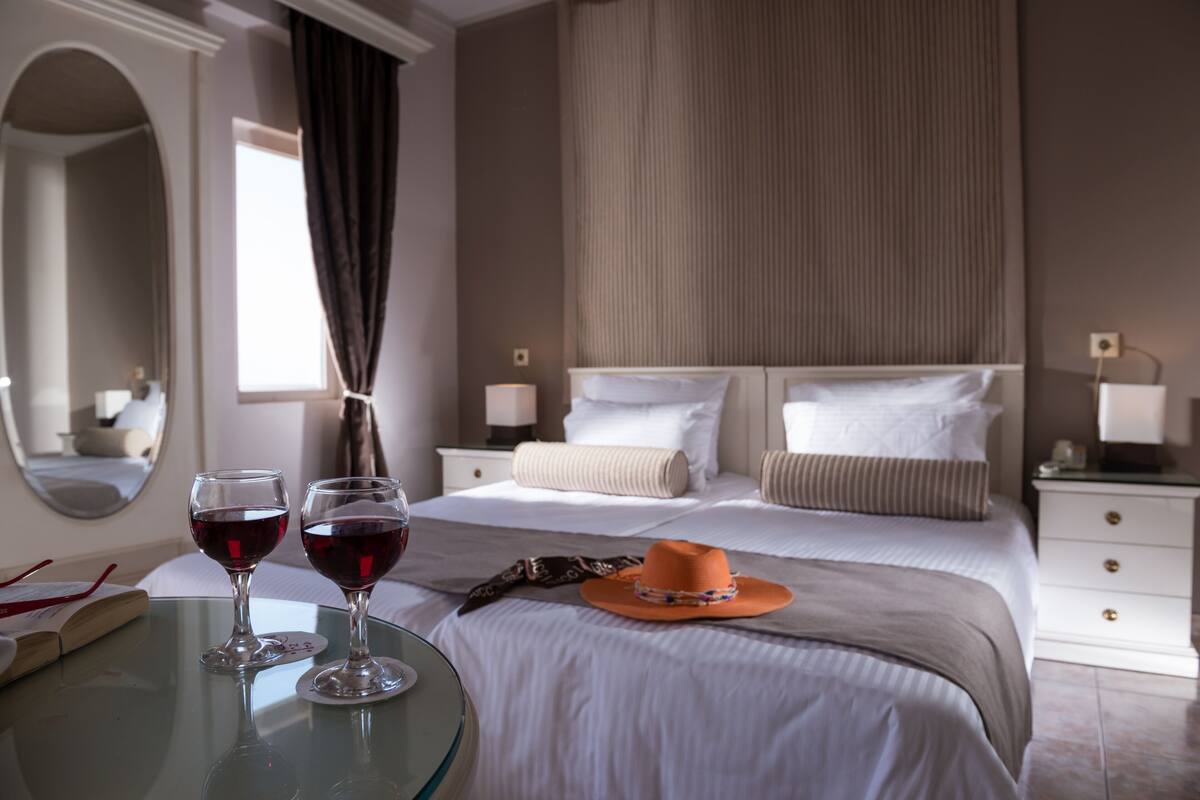 A hotel room with a bed, two bedside tables, a nightstand, a mirror, and two wine glasses on a table.
