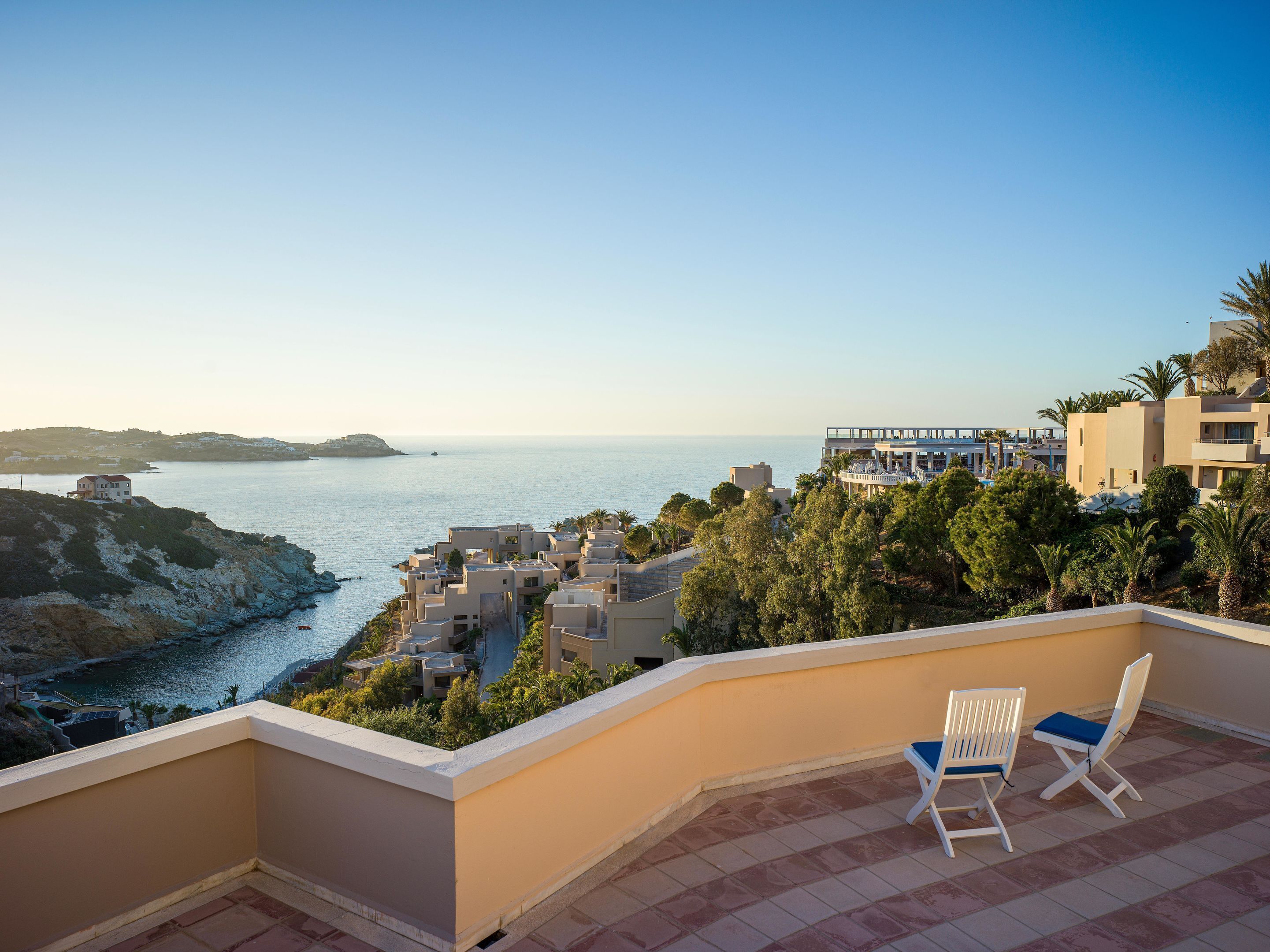 A balcony with two white chairs and a blue cushion, overlooking a coastal town with buildings and a clear sky.