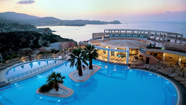 A large, circular swimming pool with palm trees and lounge chairs, surrounded by a building with multiple balconies and a view of the sea. - Athina Palace Resort & Spa (Malevizi)