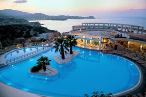 Une grande piscine circulaire, bordée de palmiers et de chaises longues, entourée d’un bâtiment doté de nombreux balcons et offrant une vue sur la mer.