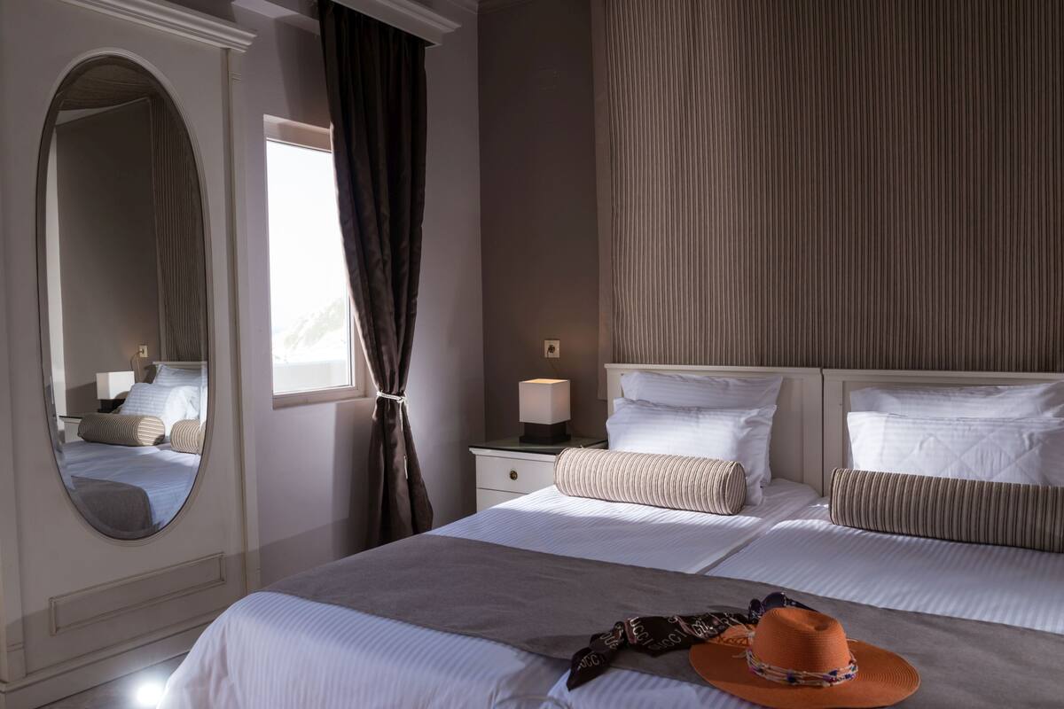 A hotel room with a large bed, a bedside table with a lamp, a mirror, and a window with curtains.