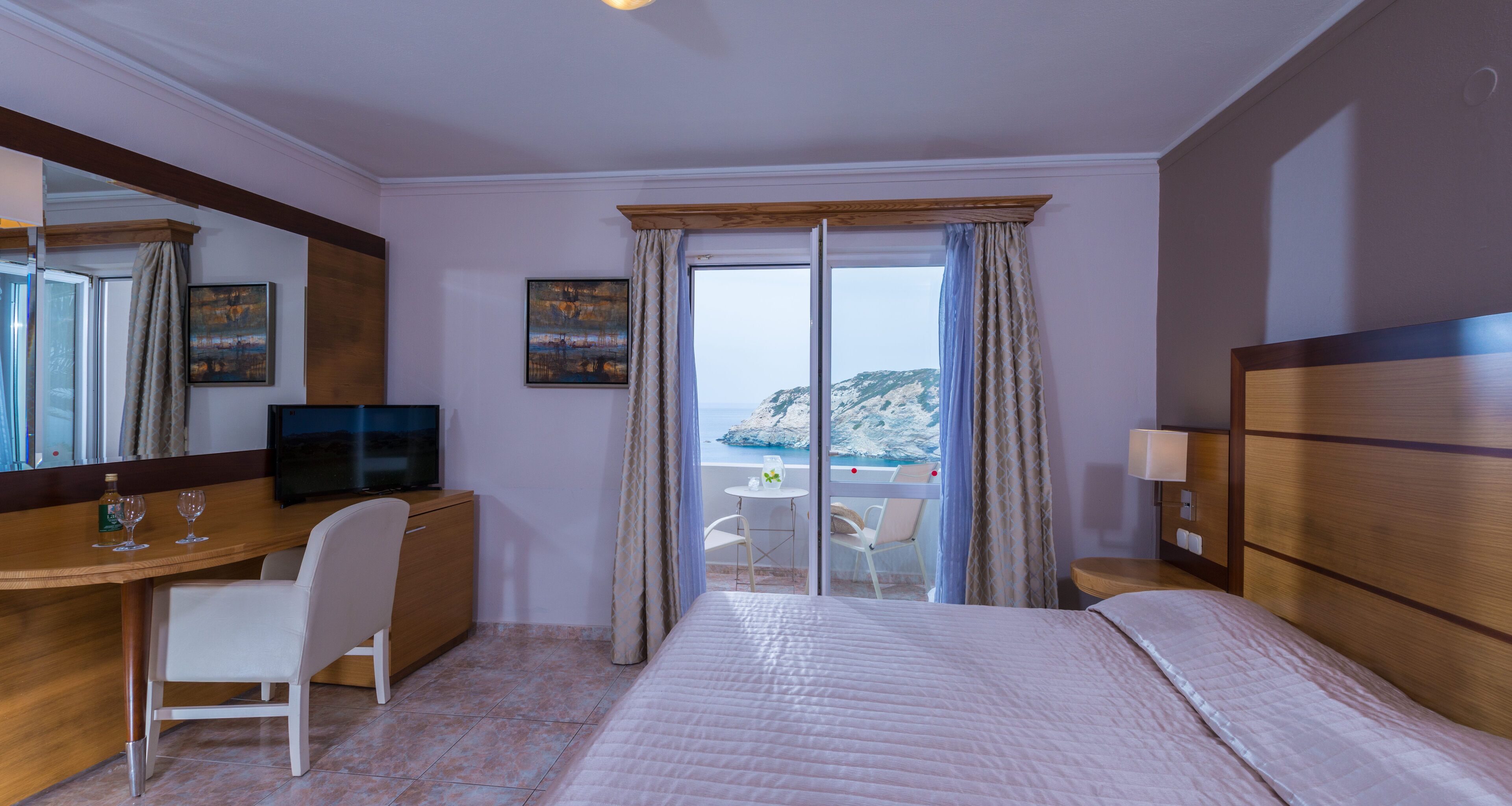 A hotel room with a bed, a desk with a chair, a television, and a view of the sea through a sliding door.