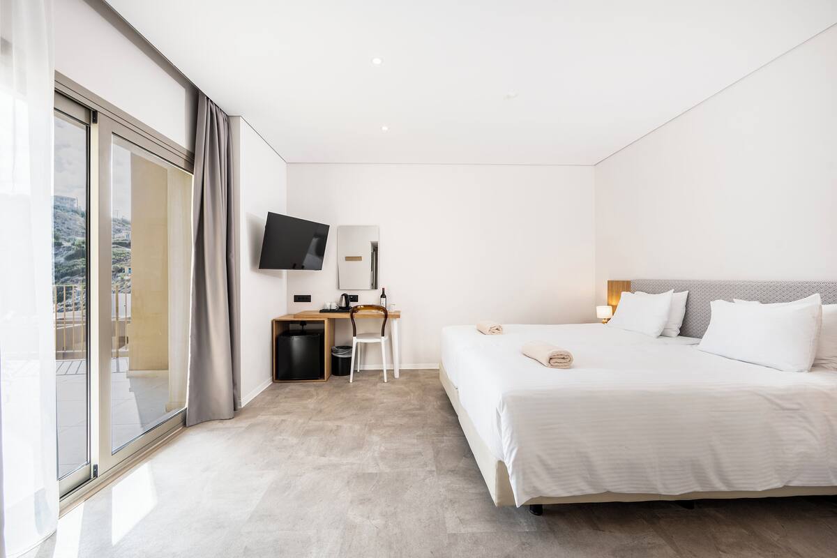 A modern hotel room with a large bed, a desk with a chair, a television, and a sliding glass door to the outside.