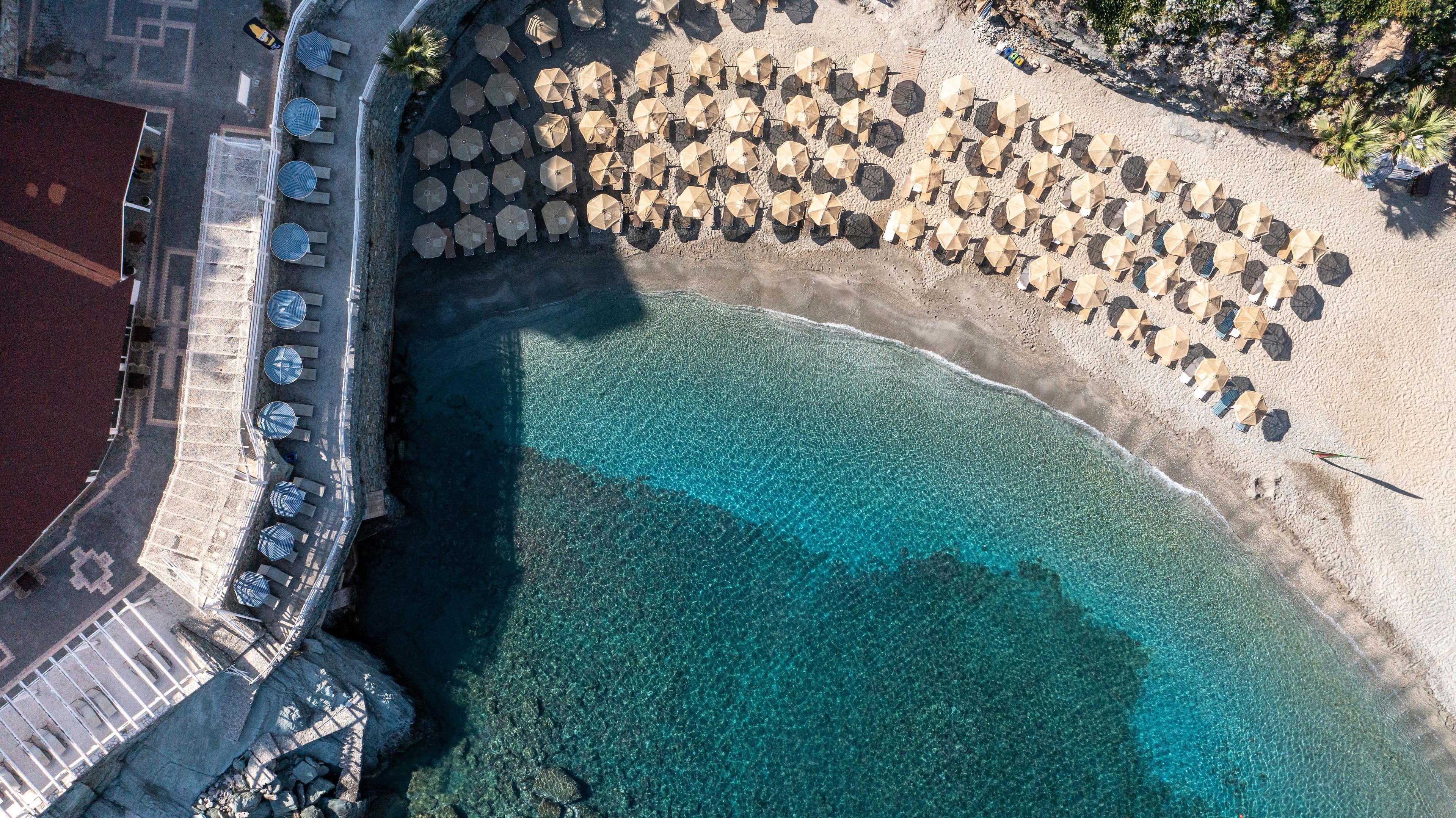 An aerial view of a beach with rows of umbrellas and lounge chairs, a clear blue sea, and a rocky shoreline.