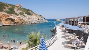 A coastal beach scene with a rocky hillside, clear blue water, and a row of beach chairs and umbrellas. - Athina Palace Resort & Spa (Malevizi)