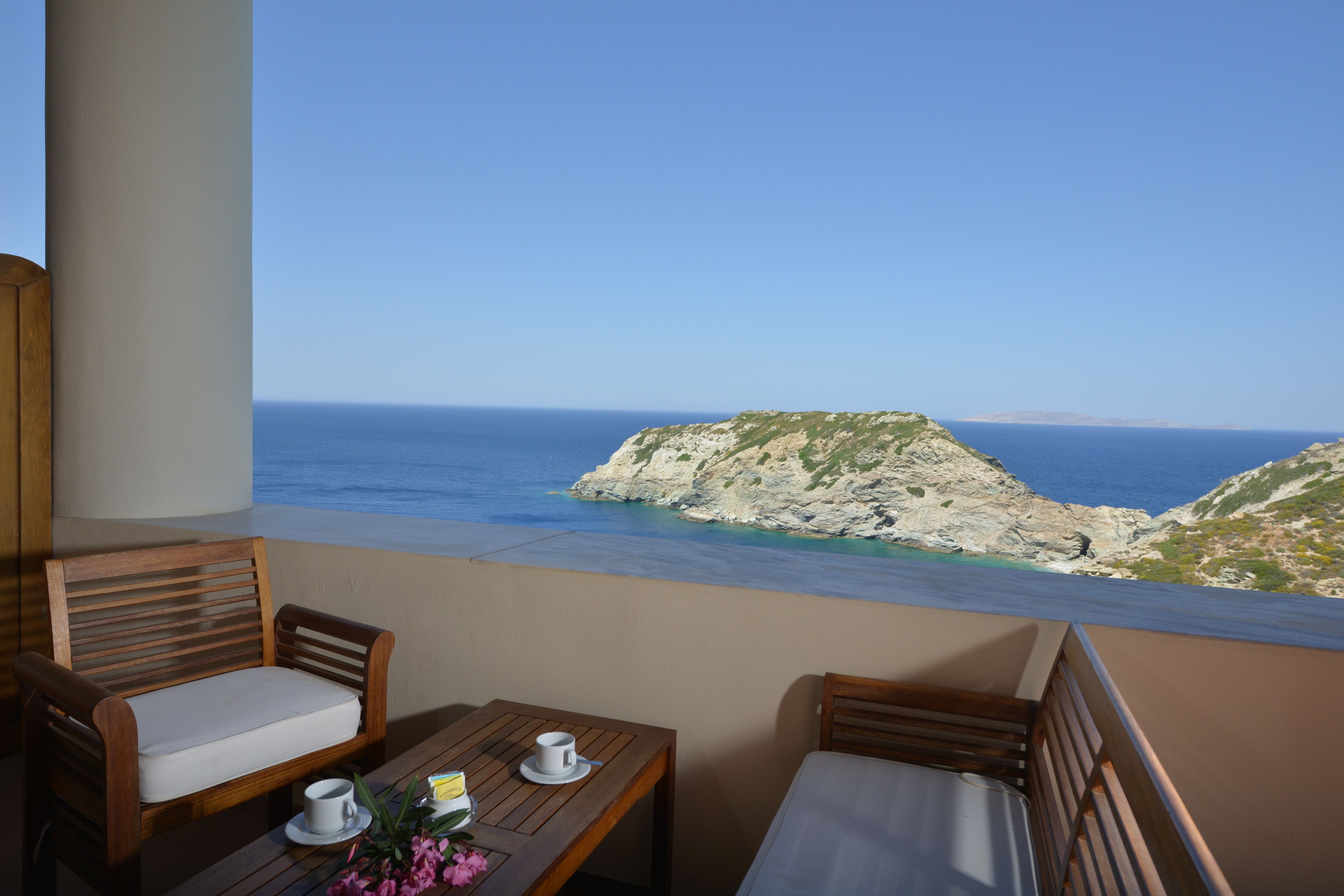 A balcony with wooden furniture, a table with a cup and flowers, and a view of the sea and rocky landscape.