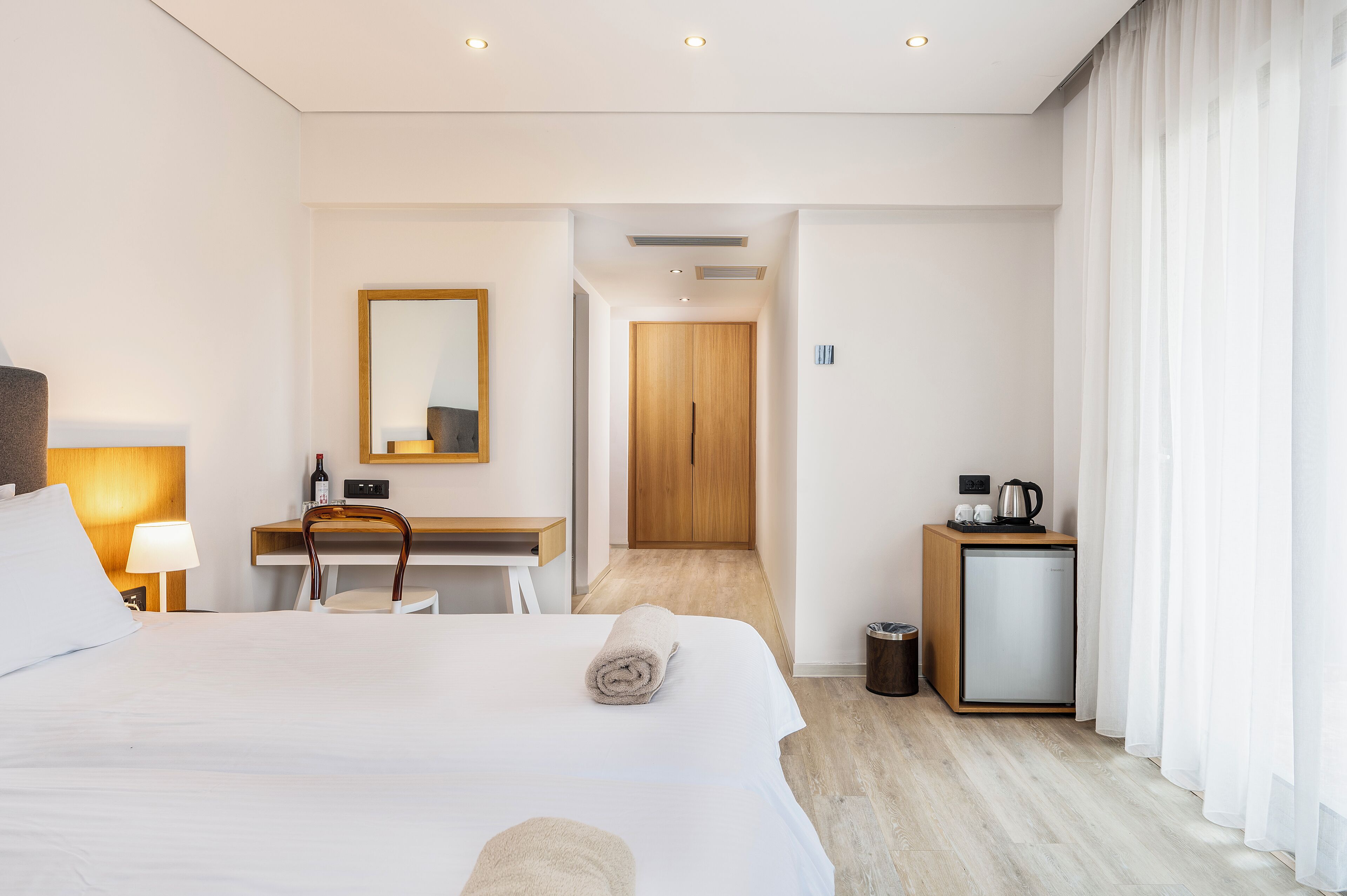 A hotel room with a bed, a desk with a chair, a small kitchenette, a mirror, and a door leading to another room.