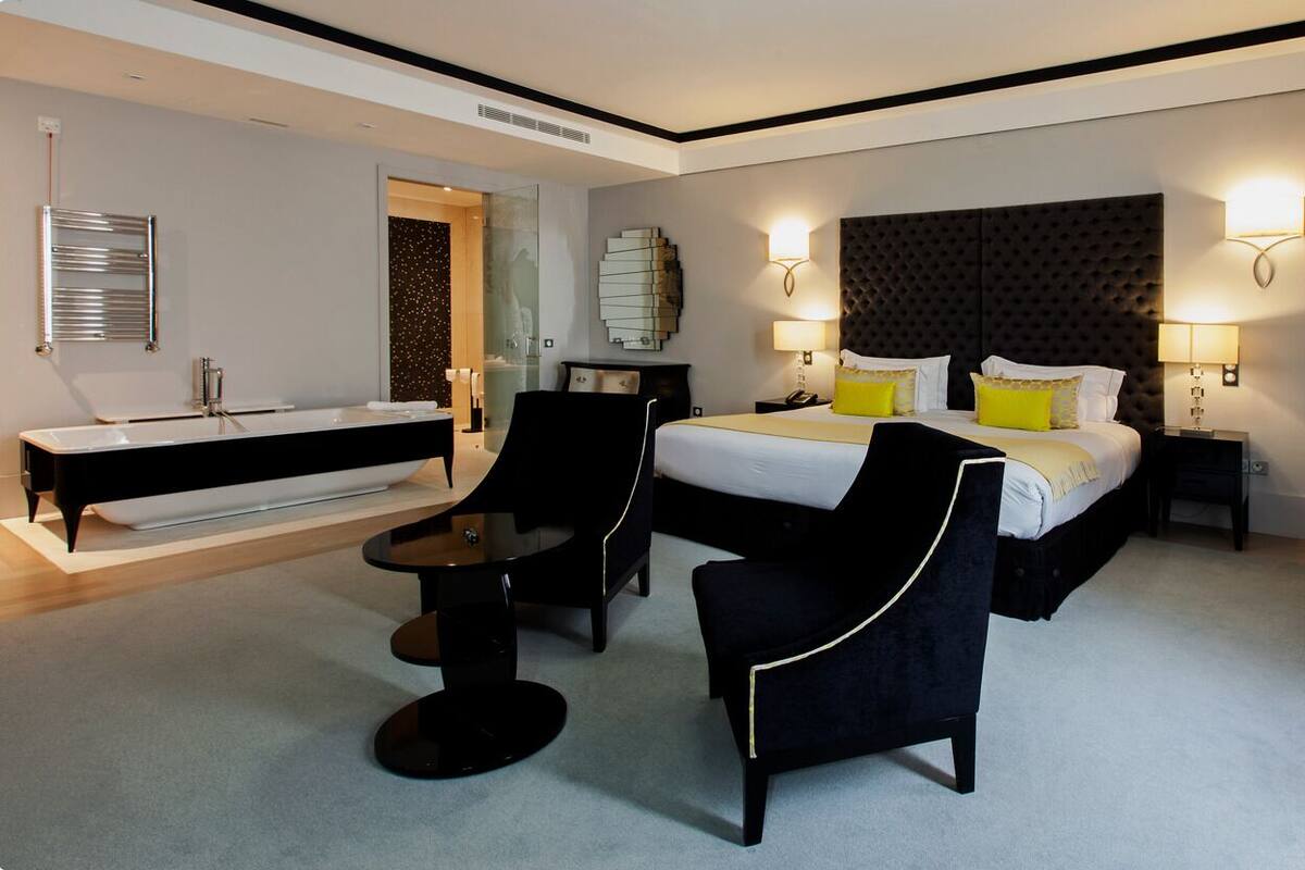 A modern hotel room with a large bed, bedside lamps, a vanity with a mirror, and a seating area with a table and chairs.