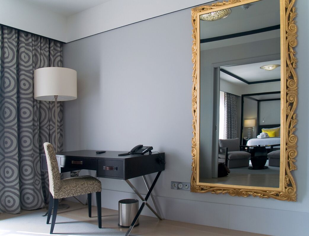 A hotel room with a desk, chair, and a large ornate mirror.
