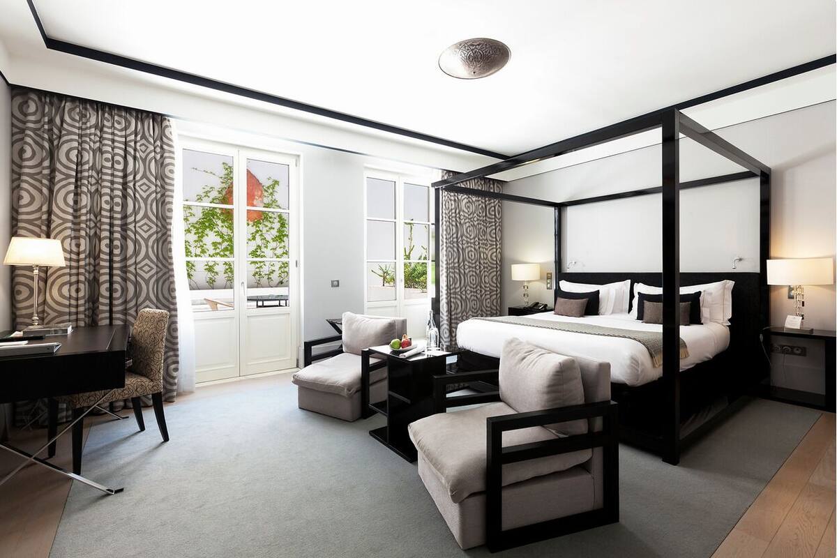 A modern hotel room with a four-poster bed, a desk with a chair, a small table with a lamp, and a view of greenery outside.