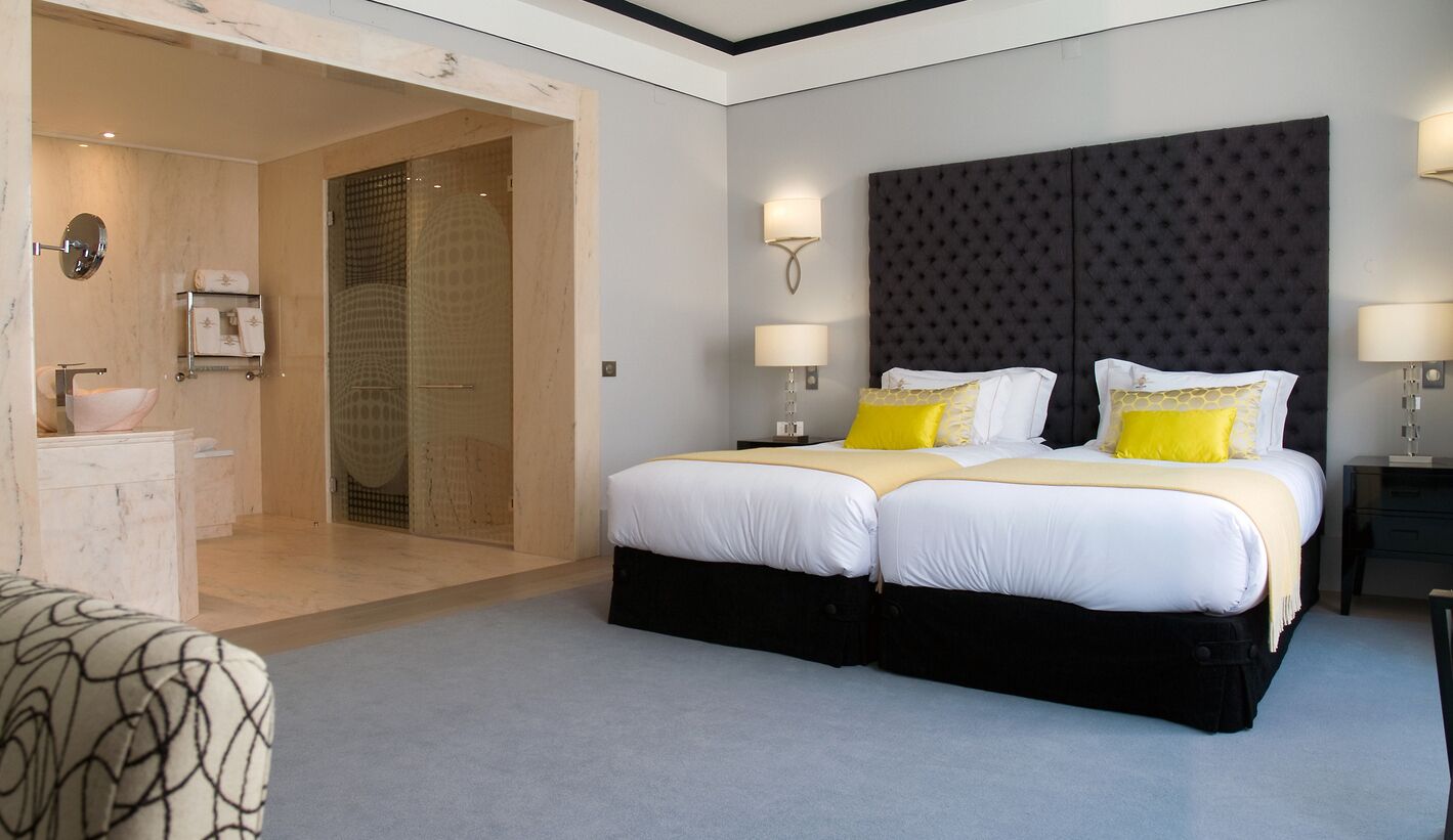A double bed with white and yellow bedding, a dark tufted headboard, two bedside lamps, and a small bedside table.