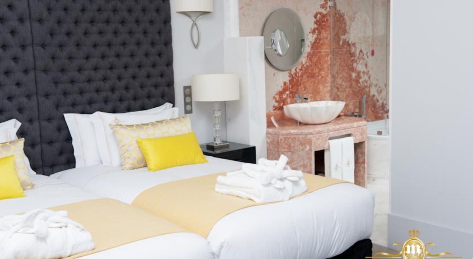 Habitación de hotel con una cama grande, una mesita de noche con lámpara, un espejo en la pared y un baño con lavamanos y un espejo amplio.