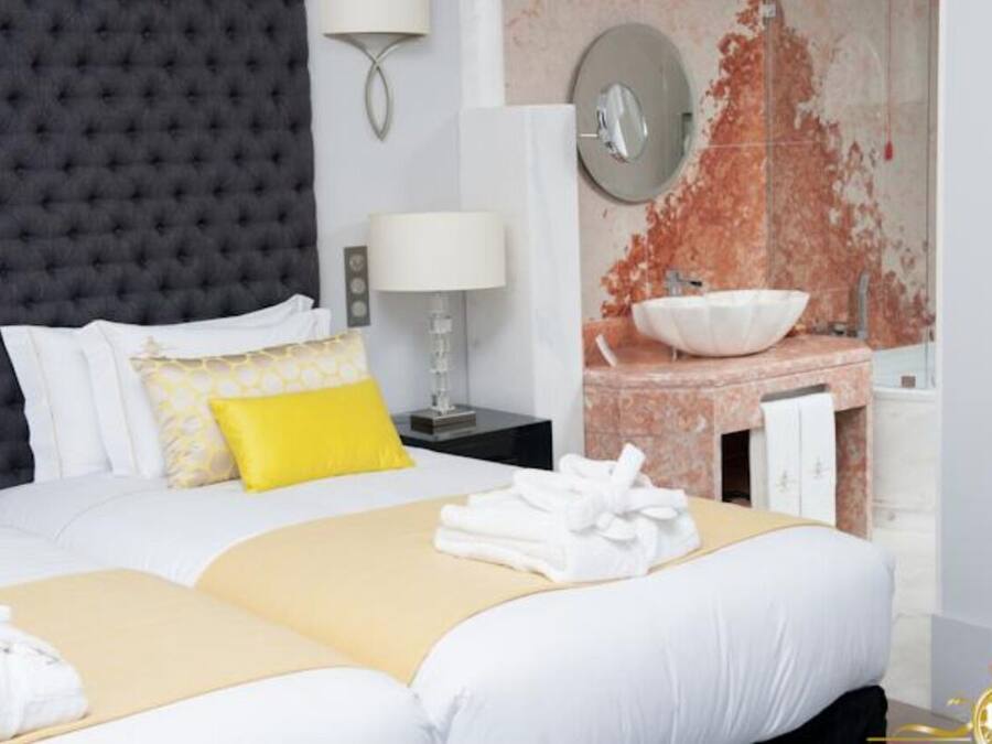 Habitación de hotel con una cama grande, una mesita de noche con lámpara, un espejo en la pared y un baño con lavamanos y un espejo amplio.