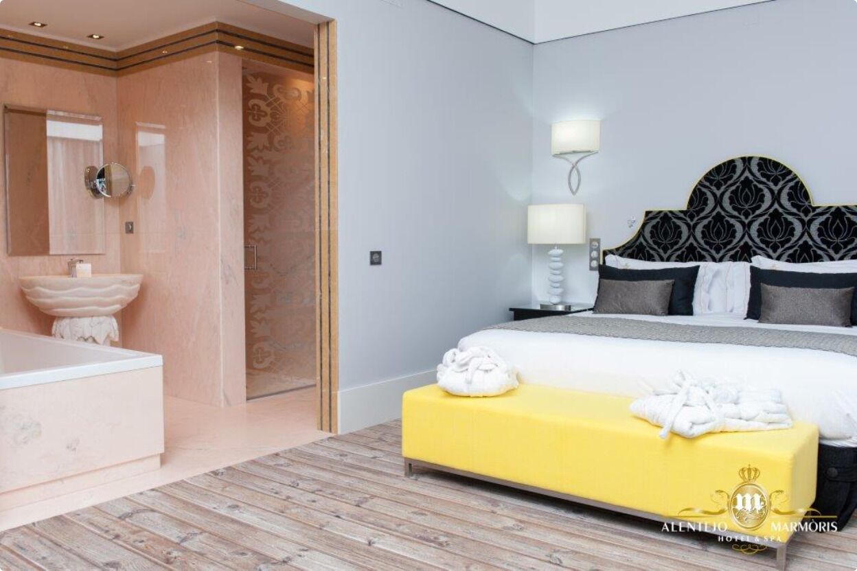 A modern hotel room with a large bed, a yellow bench, a bathroom with a large sink, and a mirror.