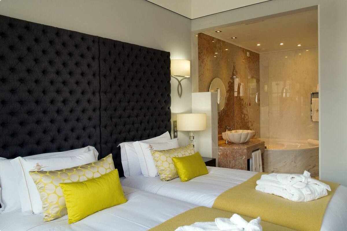 A hotel room with a tufted headboard, two beds with white and yellow bedding, a bedside table with a lamp, a bathroom with a marble sink and bathtub, and towels on the bed.