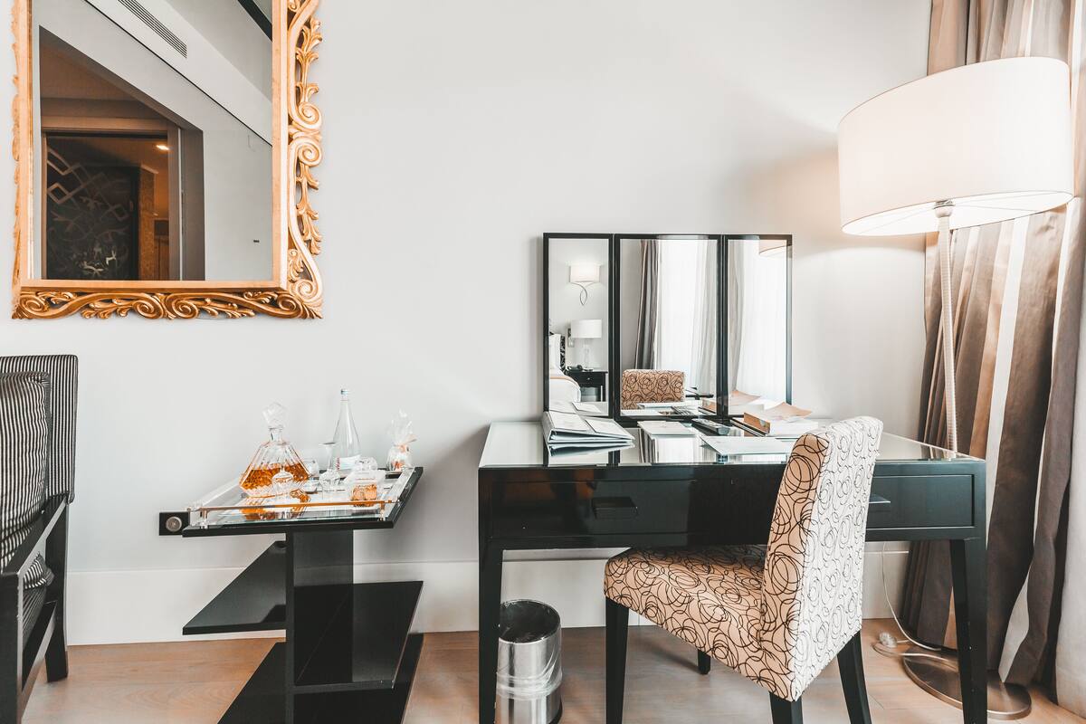 A modern hotel room with a black desk, a patterned chair, a large mirror, and a lamp.