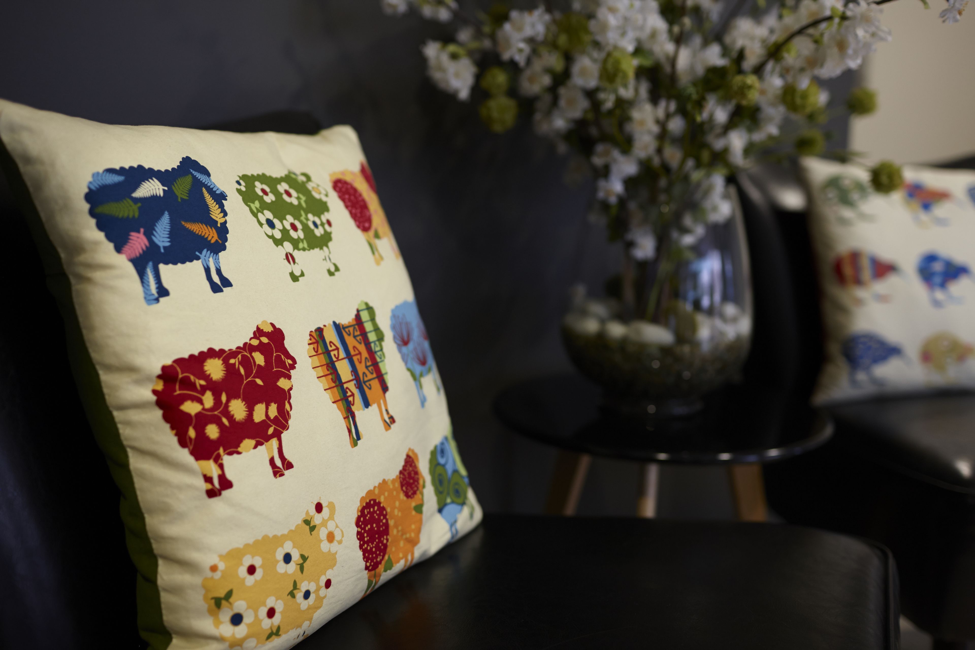 Un cojín con estampados de animales y flores coloridos, un jarrón con flores blancas y una silla de color oscuro.
