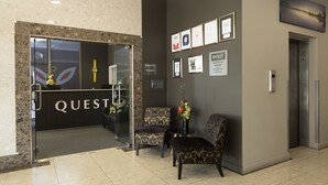 A reception area with a 'QUEST' sign, two patterned chairs, a vase with flowers, and framed certificates on the wall.