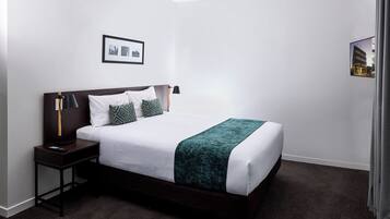 Cama bem arrumada com cabeceira escura, roupa de cama branca e manta verde. Tem uma mesa de cabeceira com abajur e um quadro emoldurado sobre a cama.