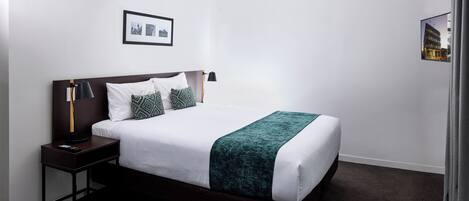 A neatly made bed with a dark headboard, white bedding, and a green throw. There is a nightstand with a lamp and a framed picture above the bed.
