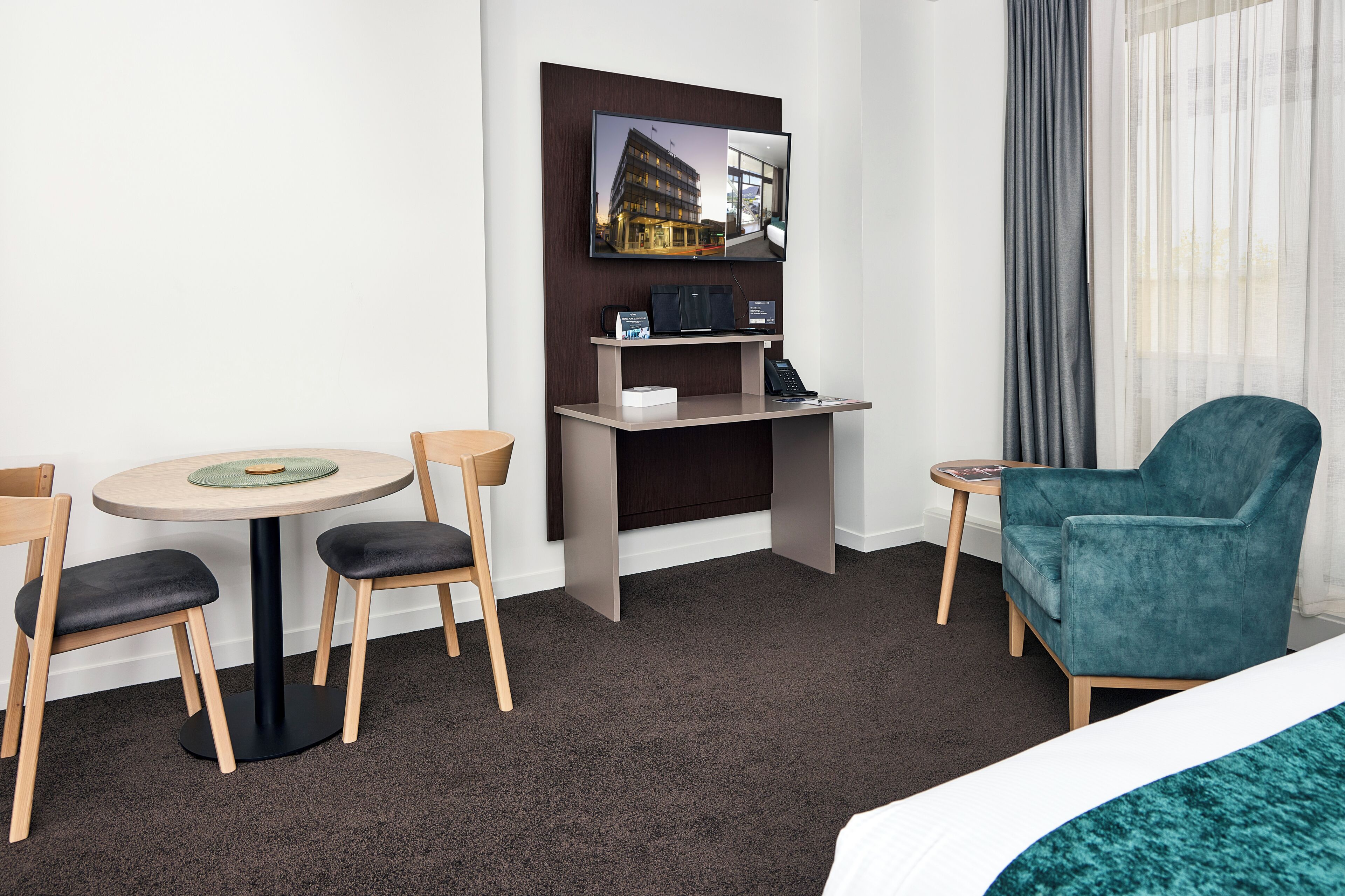 A hotel room with a bed, a desk with a computer, a chair, a small table, and a view of a building through the window.