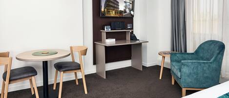 A hotel room with a bed, a desk with a computer, a chair, a small table, and a view of a building through the window.