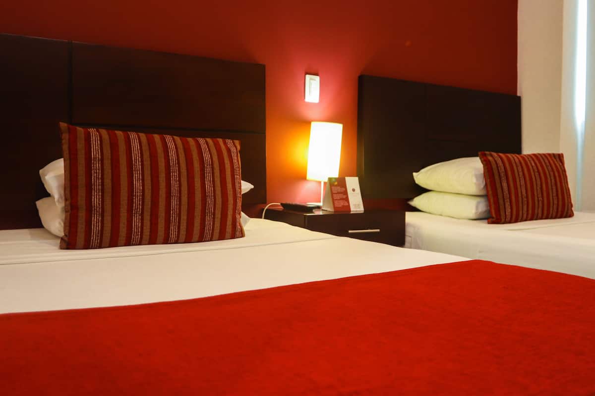 A hotel room with two beds, each with striped pillows, a red blanket, and a bedside table with a lamp and a phone.