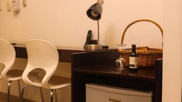 A hotel room with a flat-screen TV mounted on the wall, a desk with a lamp, a small refrigerator, and two white chairs.