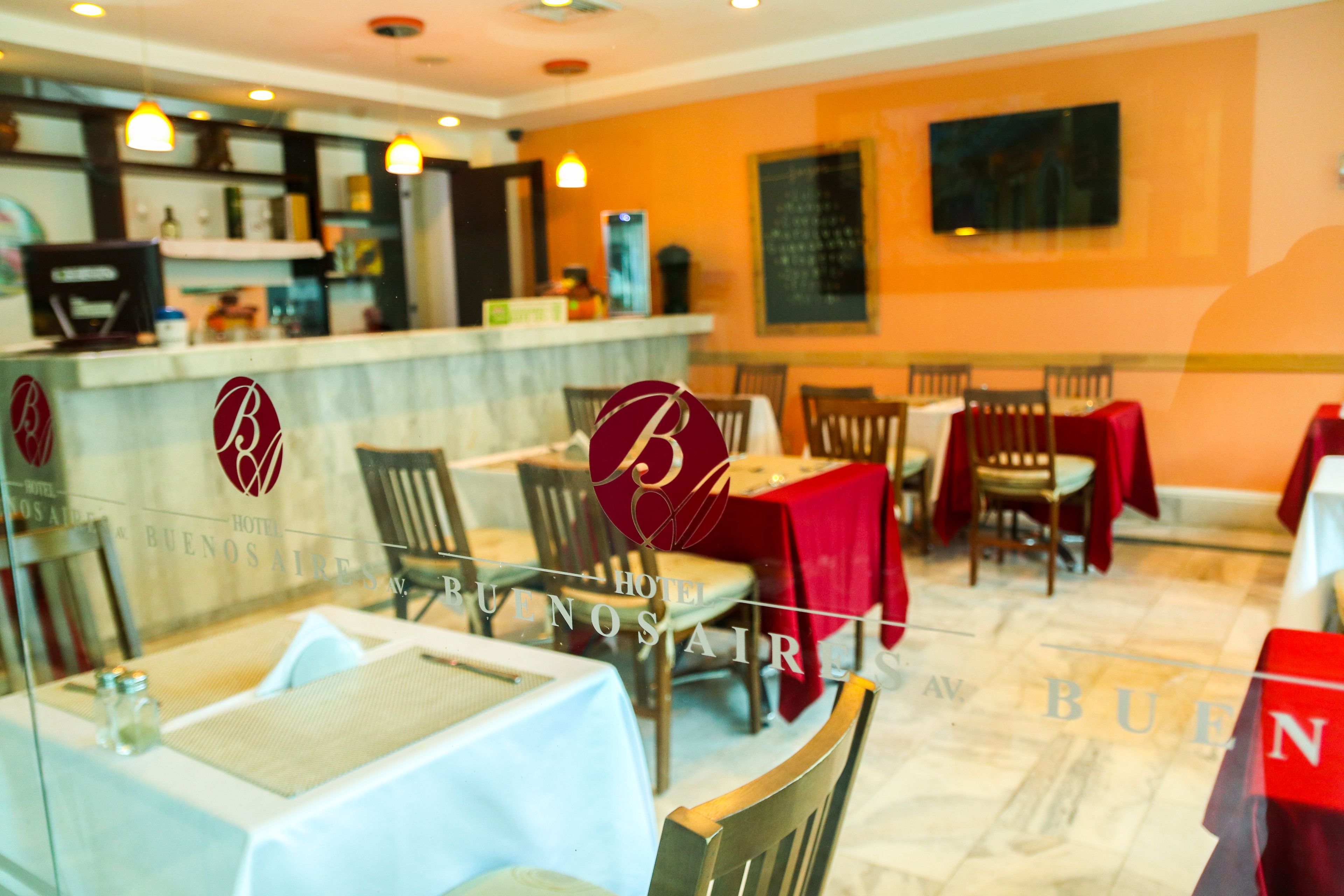 A restaurant with tables covered in white tablecloths, wooden chairs, and red tablecloths. The restaurant has a counter with a menu and a television screen. The walls are orange, and there is a chalkboard on the wall.