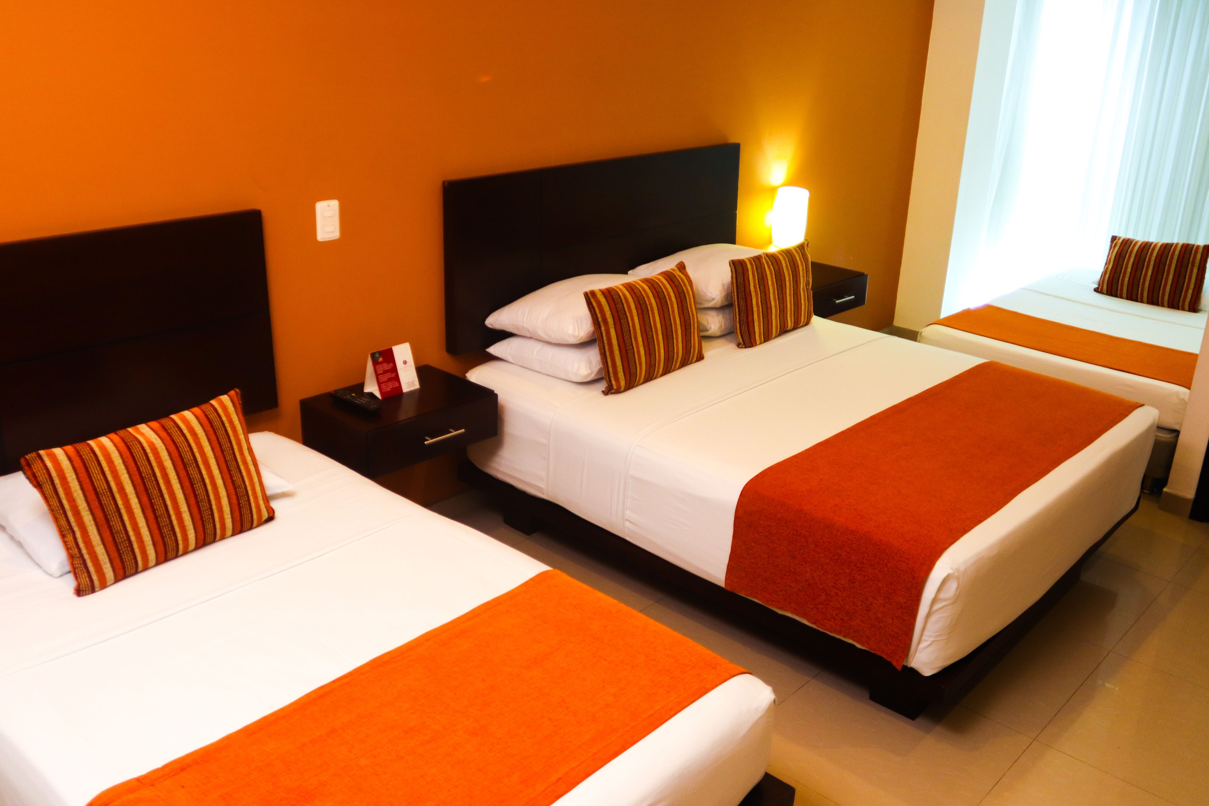 A hotel room with two beds, each with a striped pillow and a red blanket. There is a nightstand with a lamp between the beds.