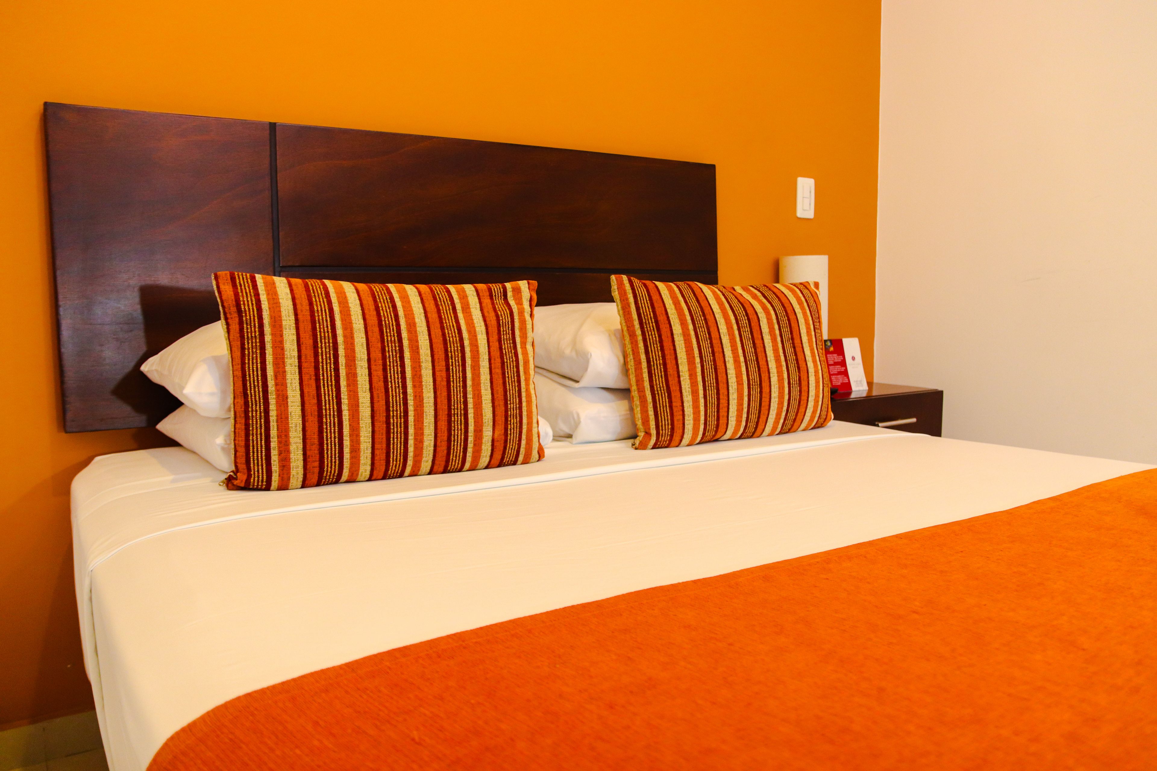 A neatly made bed with striped pillows and a white comforter, set against an orange wall.