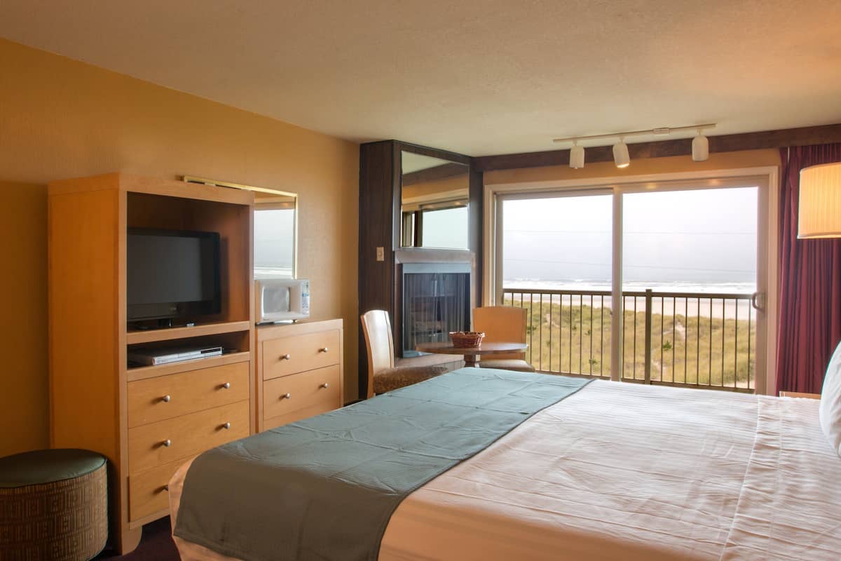 A hotel room with a bed, a television, a desk, and a window with a view of the beach.
