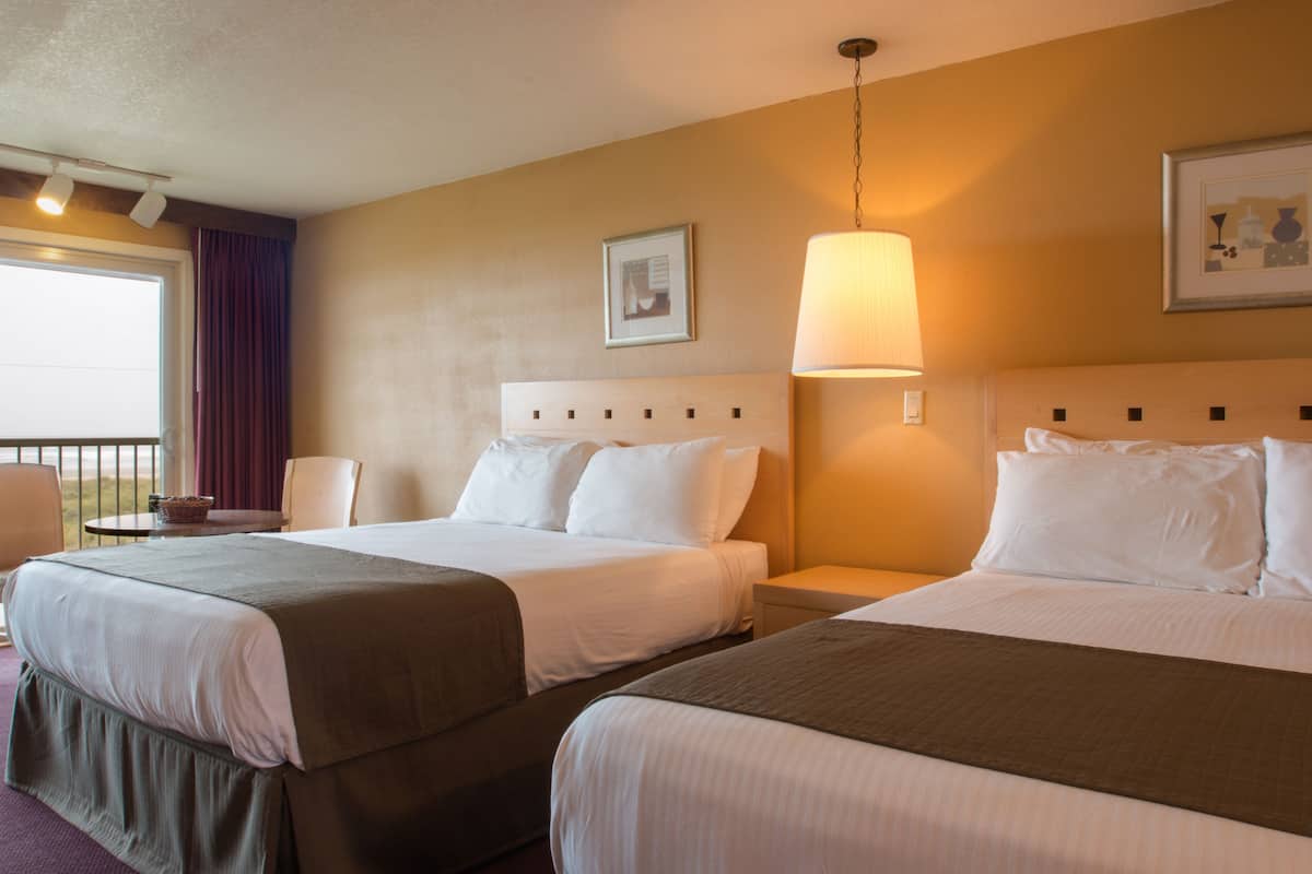 A hotel room with two beds, a bedside table, a lamp, a chair, and a window with curtains.