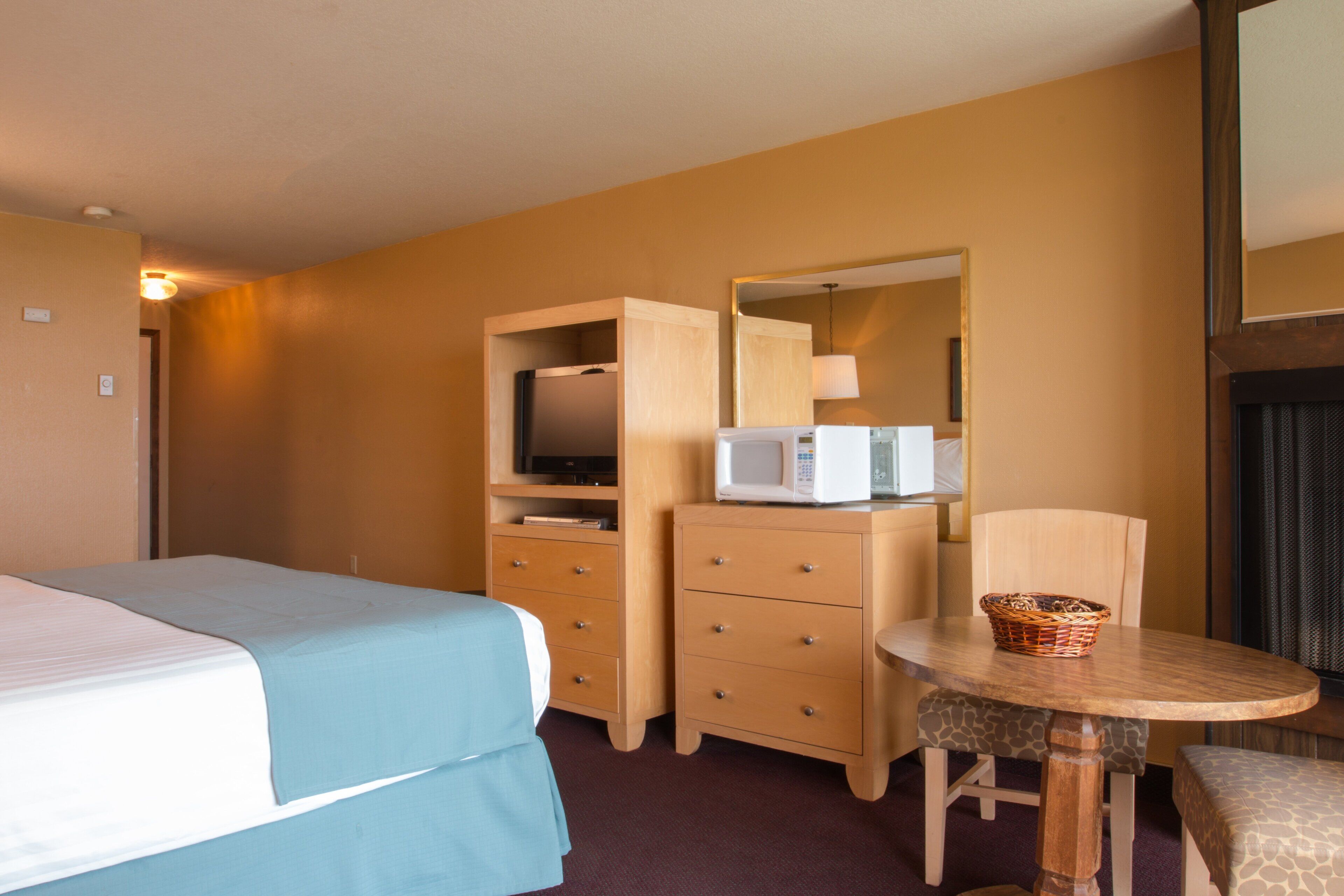 A hotel room with a bed, a wooden dresser with a microwave, a small table with a basket, and a chair.
