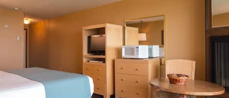 A hotel room with a bed, a wooden dresser with a microwave, a small table with a basket, and a chair.