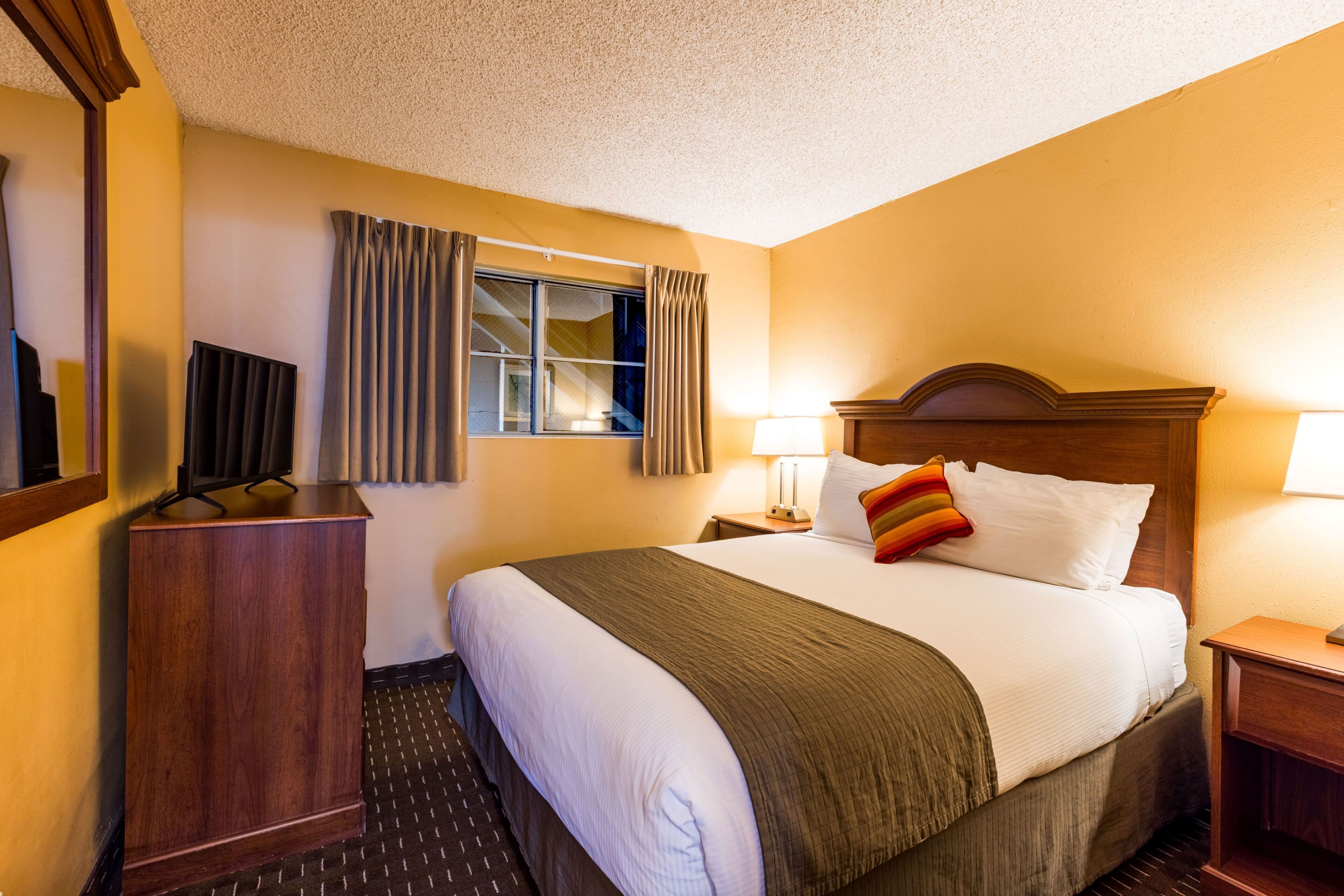 A hotel room with a large bed, a wooden headboard, a nightstand with a lamp, a desk with a television, and a window with curtains.