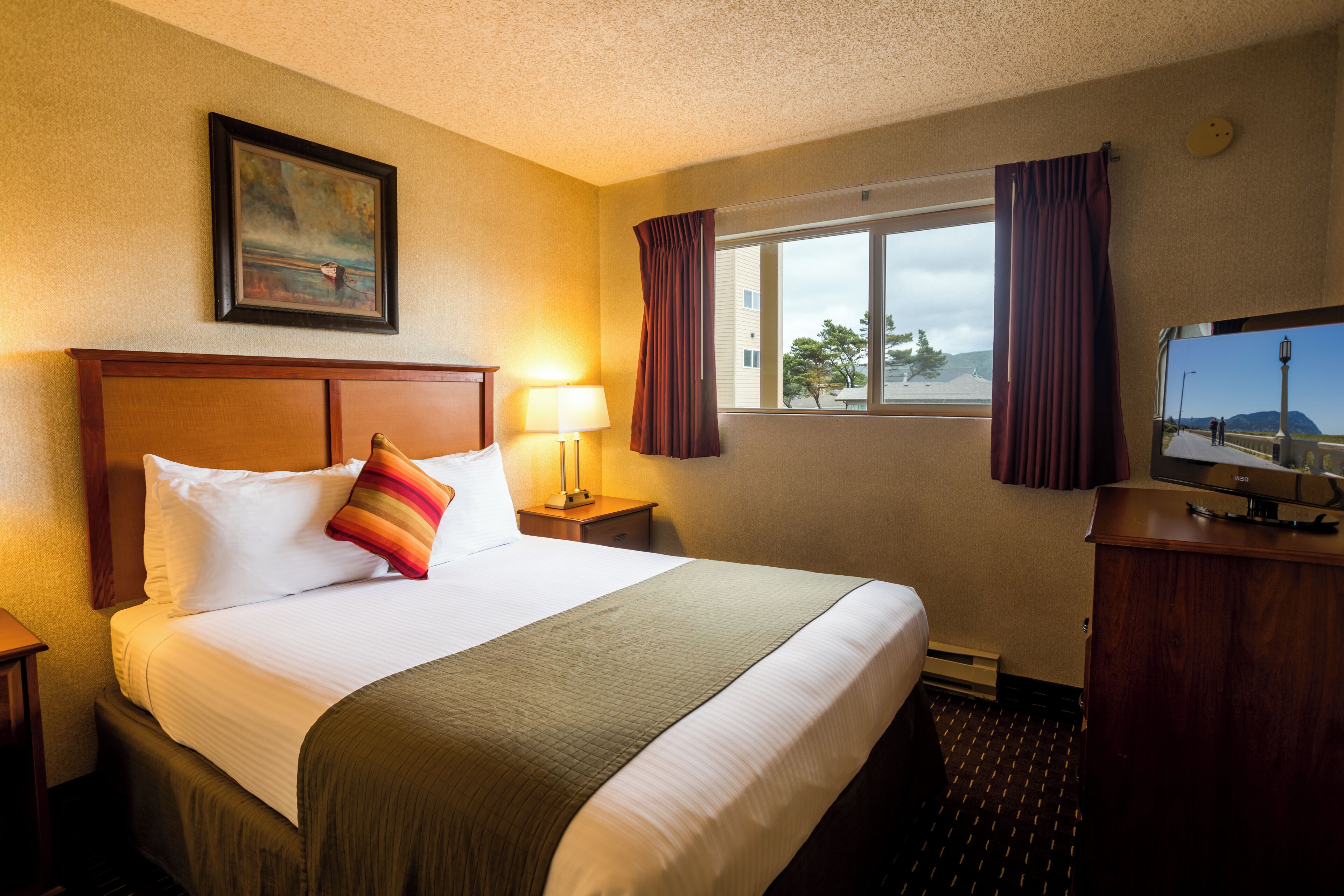 A hotel room with a large bed, a nightstand with a lamp, a TV on a wooden cabinet, a window with curtains, and a painting on the wall.