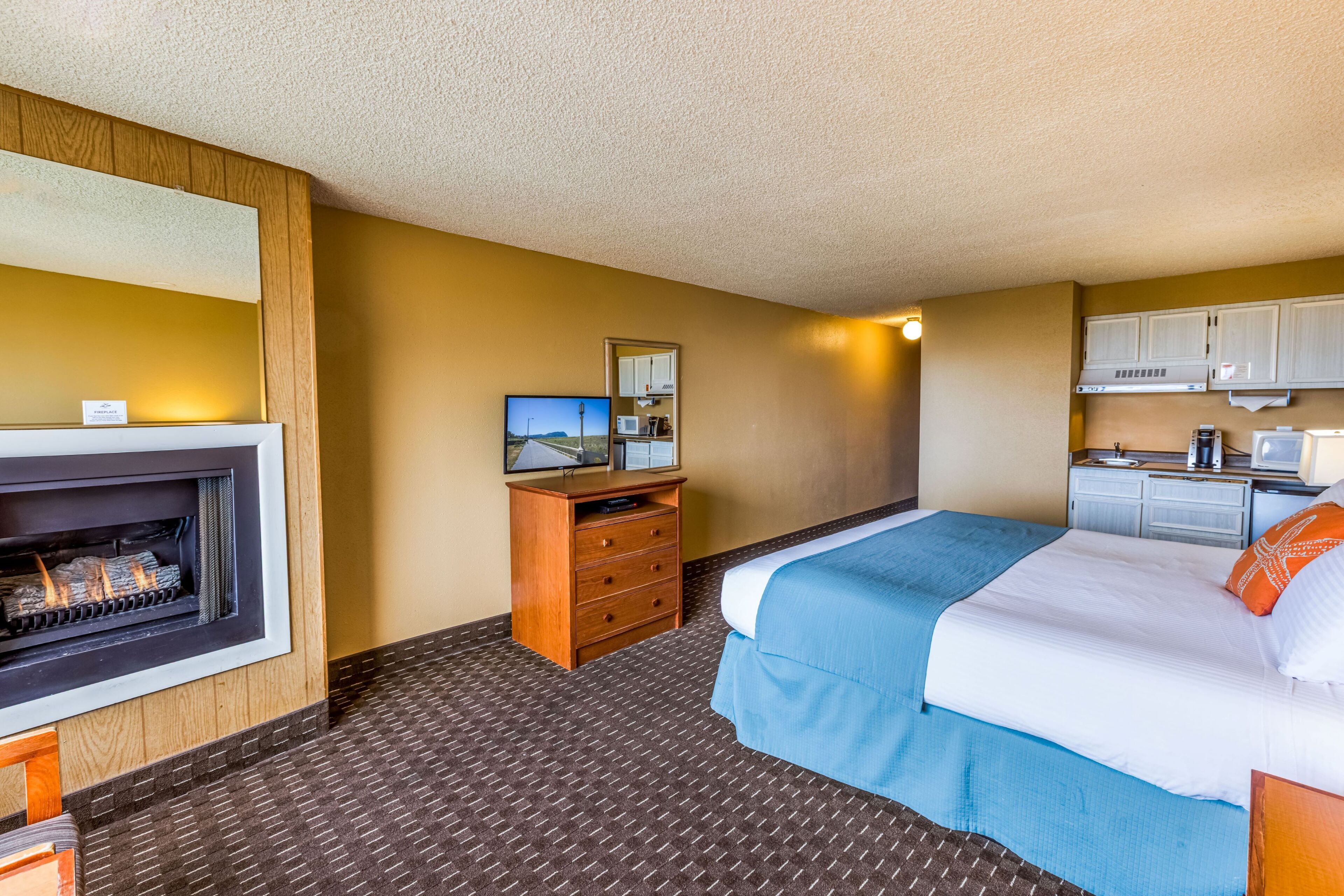 Een hotelkamer met een open haard, een bed met blauw beddengoed, een houten dressoir, een televisietoestel en een kitchenette.