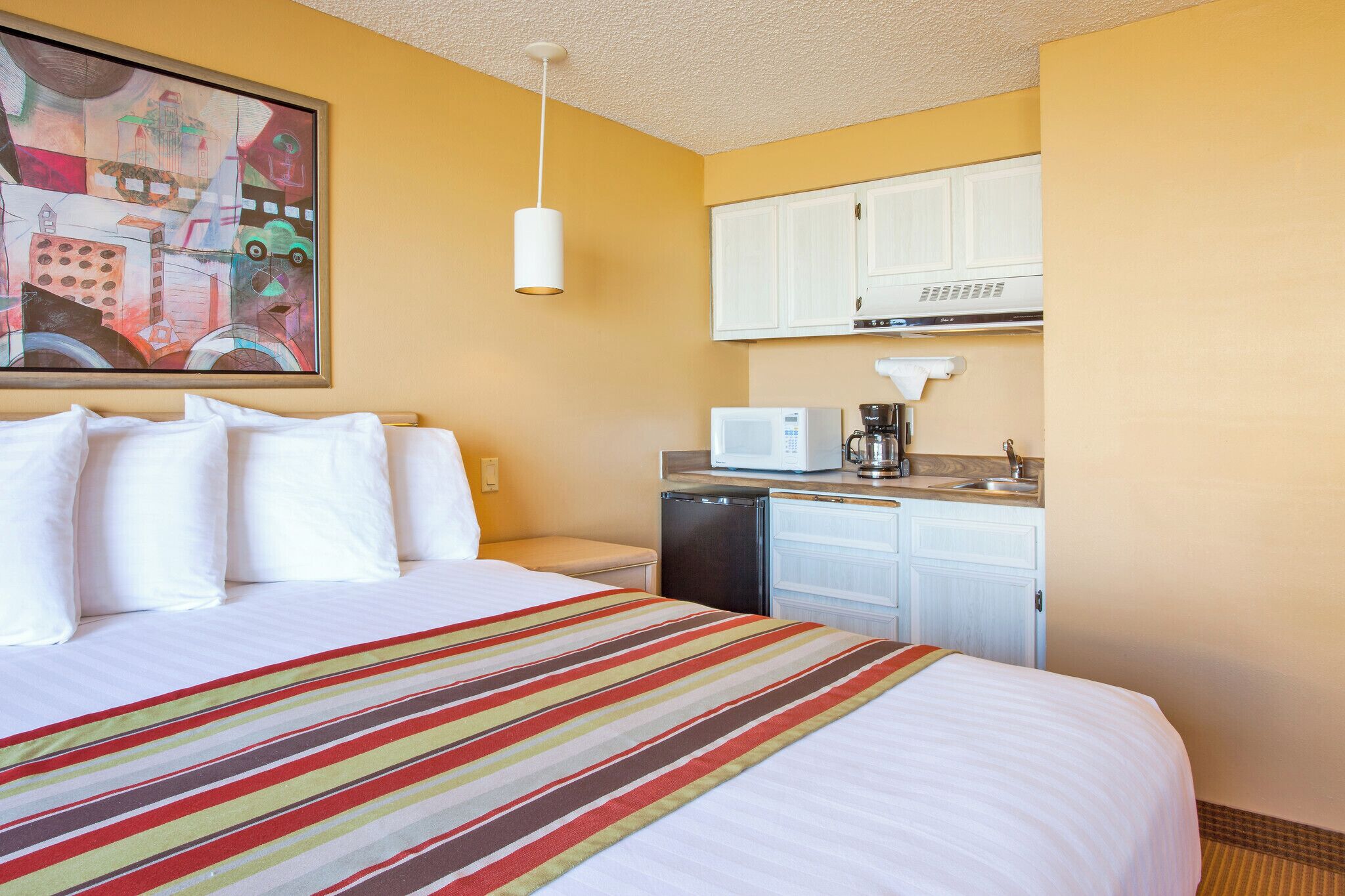 A hotel room with a bed, a kitchen area with a microwave, a coffee maker, and a dishwasher.