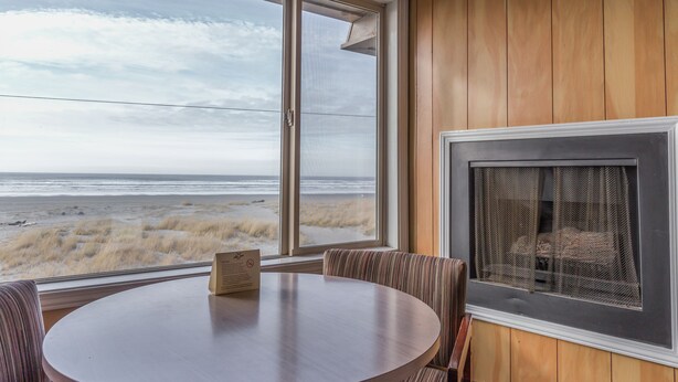 A room with a large window overlooking the beach, a round table, and a fireplace. - Ebb Tide Oceanfront Inn (Seaside)