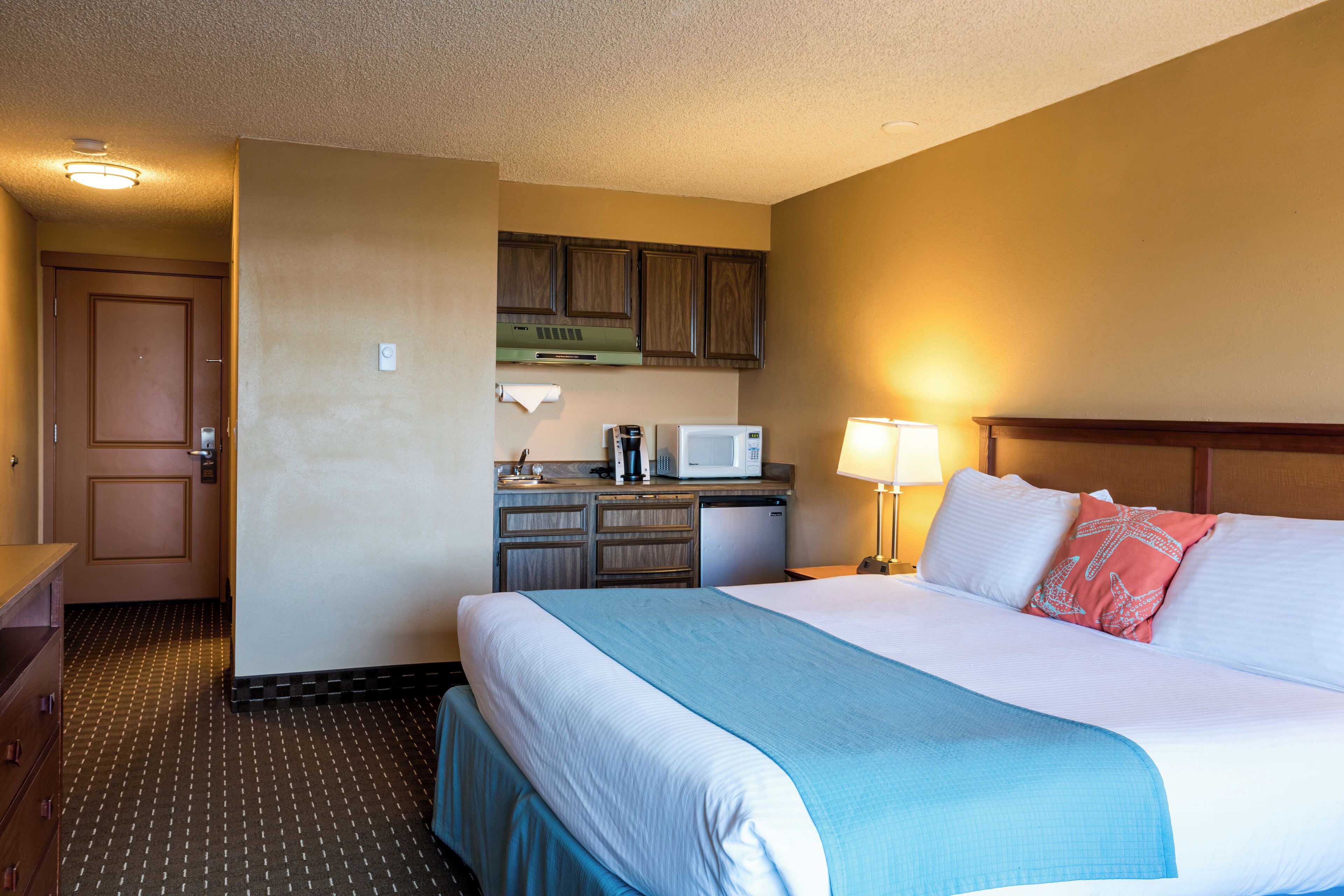 A hotel room with a bed, a desk with a microwave, a coffee maker, and a small kitchenette.